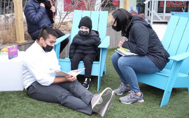 Creating Healing Spaces at our March Wellbeing Jamboree 2 Adult with a book sits on grass, while child and two other adults look on.