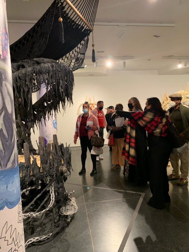 Curators for Educators: Health + Wellbeing are Human Rights 3 Eight people stand in an open gallery while one wearing a plaid shawl points to a mixed media sculpture.