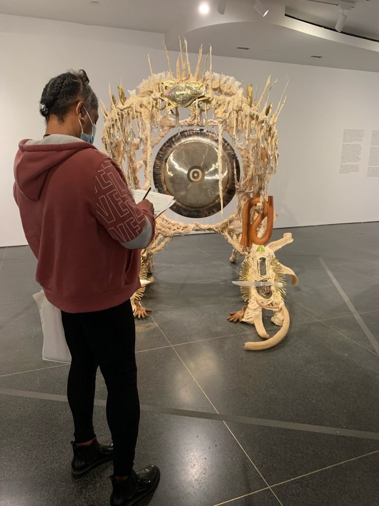 Curators for Educators: Health + Wellbeing are Human Rights 2 One educator stands with their back towards the camera as they take notes while viewing a mixed media artwork that includes a healing gong at the center.