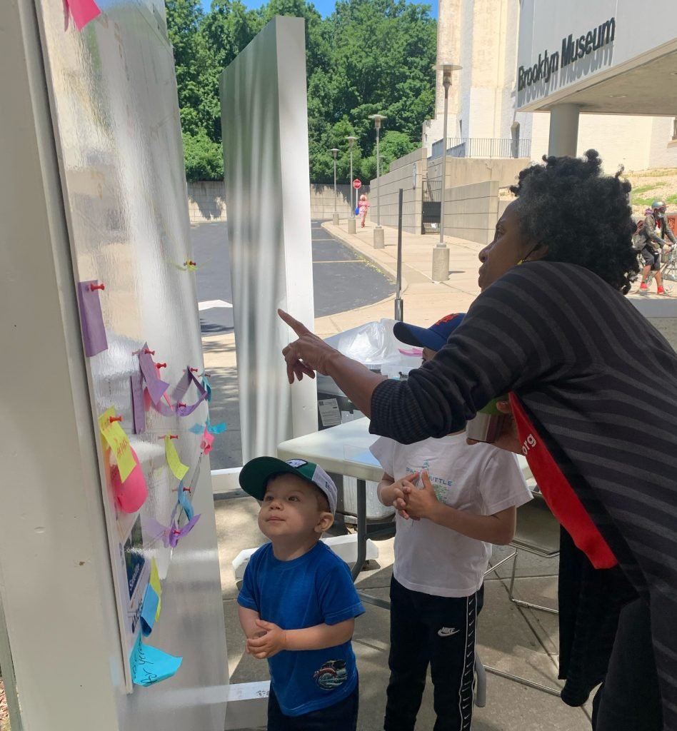 Bring the Cool! Honoring Juneteenth with Brooklyn Museum 3 Two children look on as a Cool Culture staff member points to a movable wall panel with post-it notes and a sun glare that obscures a map.