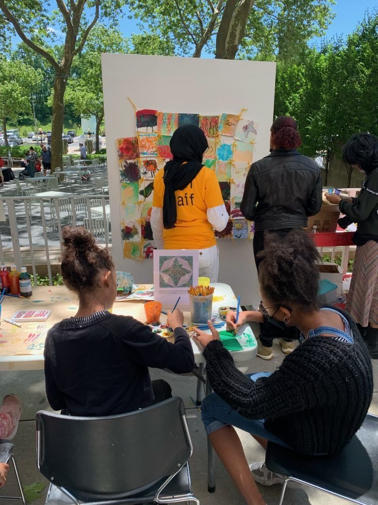 Bring the Cool! Honoring Juneteenth with Brooklyn Museum 1 Two individuals stand with their backs to the camera facing a movable wall panel with a quilt-like mural made of painted squares while two other individuals sit at a table painting in the foreground.