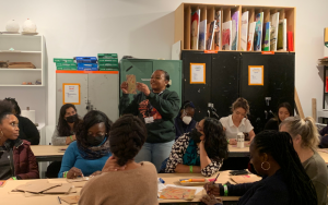 Museum staff holds up example of art in between tables of educators working on their own art. The room has shelves filled with paper.